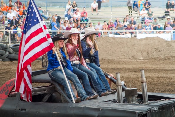 Rodeo queens riding into an arena on a demolition derby car, holding an American flag