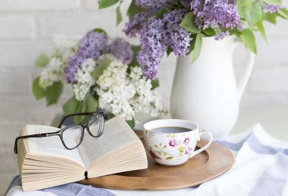 Open book sits on a tray next to a mug of cocoa and a vase of lilac flowers
